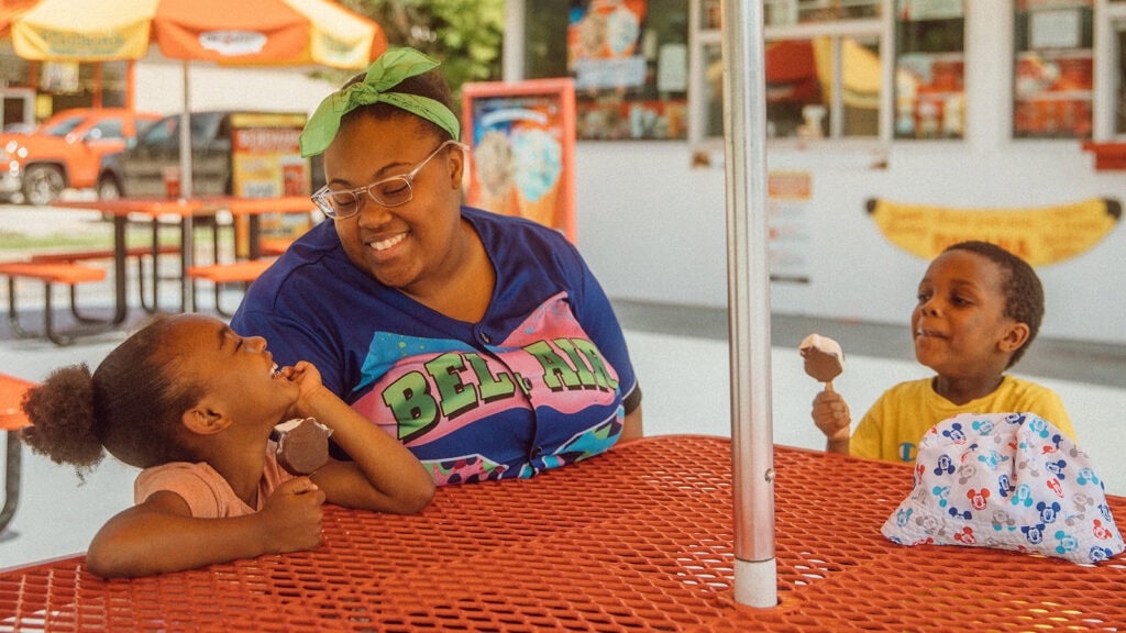A mom and two kids eat ice cream under an umbrella in the summer heat.